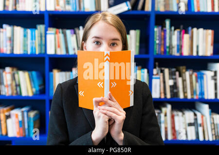 Junges Mädchen versteckt sich hinter Buch in einer öffentlichen Bibliothek Stockfoto