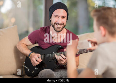 Junger Mann zu Hause auf der Couch sitzen, Gitarre spielen für Teenager, die ein Handy Bild Stockfoto