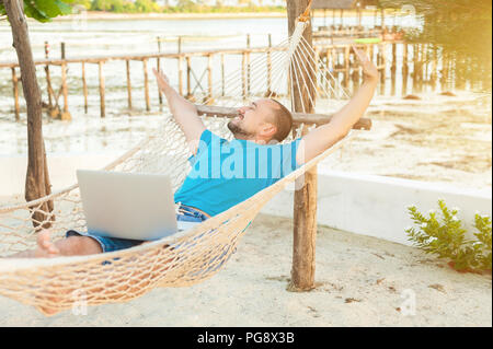 Der junge Mann liegt in einer Hängematte und ist mit einer erfolgreichen Transaktion zufrieden. Das Konzept der Fernbedienung. Stockfoto