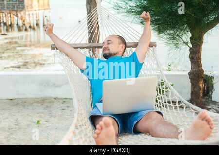 Der junge Mann liegt in einer Hängematte und ist mit einer erfolgreichen Transaktion zufrieden. Das Konzept der Fernbedienung. Stockfoto