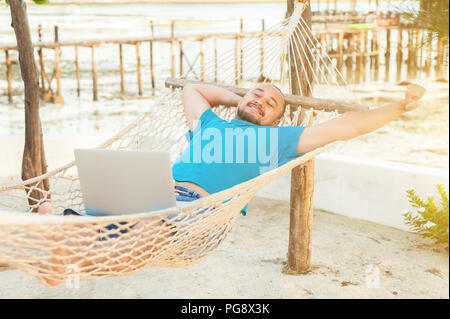 Der junge Mann liegt in einer Hängematte und ist mit einer erfolgreichen Transaktion zufrieden. Das Konzept der Fernbedienung. Stockfoto