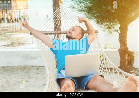 Der junge Mann liegt in einer Hängematte und ist mit einer erfolgreichen Transaktion zufrieden. Das Konzept der Fernbedienung. Stockfoto