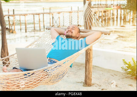 Der junge Mann liegt in einer Hängematte und ist mit einer erfolgreichen Transaktion zufrieden. Das Konzept der Fernbedienung. Stockfoto