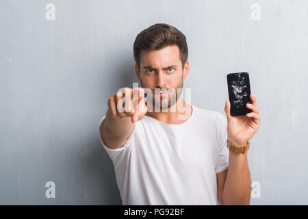 Hübscher junger Mann über Grau grunge Wand gebrochen zeigt der Bildschirm des Smartphones zeigt mit dem Finger auf die Kamera und an Sie, Hand, Zeichen, positiv und Konfi Stockfoto