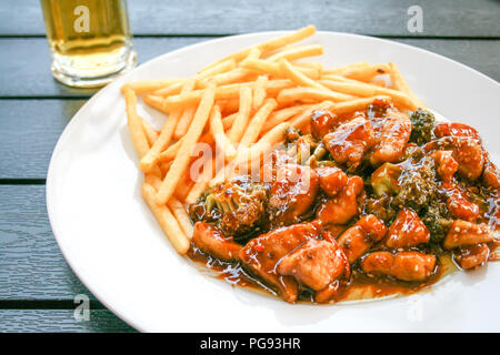 Oder im Wok gebratenen Fisch mit buntem Gemüse und Kohl, Sojasauce und Samen, Pommes frites auf einem Holztisch mit Bier im Hintergrund Stockfoto