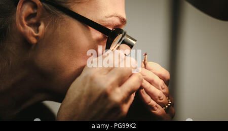 Nahaufnahme der älteren Frau goldsmith Kontrolle der Qualität eines Diamanten. Weibliche Juwelier Inspektion ein Juwel durch die Lupe in der Werkstatt. Stockfoto