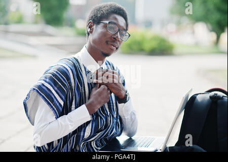 Afrikanischer mann Student in traditioneller Kleidung und Gläser mit Rucksack, Laptop im Freien arbeiten. Stockfoto