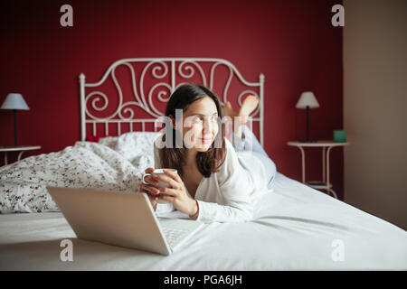 Gerne nachdenklich Frau auf dem Bett und Trinken coffrr mit Laptop zu Hause entspannen. Konzept Rest Stockfoto