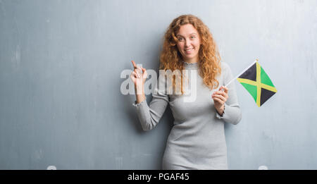 Junge rothaarige Frau über Grau grunge Wand holding Flagge von Jamaika sehr zufrieden zeigt mit Finger- und an der Seite Stockfoto