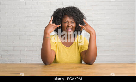 Junge afrikanische amerikanische Frau sitzt auf dem Tisch zu Hause schreien mit verrückten Ausdruck tun Rock Symbol mit hands up. Musik Star. Schwere Konzept. Stockfoto