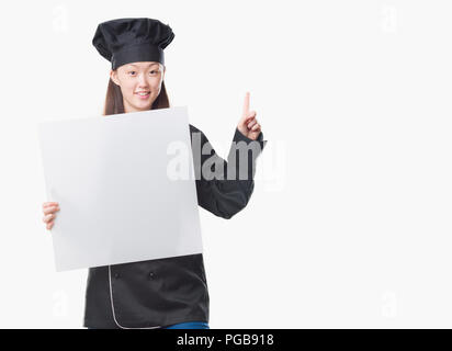 Junge chinesische Frau über isolierte Hintergrund tragen Küchenchef einheitliche Holding banner überrascht mit einer Idee oder Frage Zeigefinger mit glücklichen Gesicht, n Stockfoto