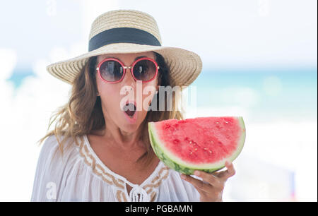 Mittleres Alter brünette Frau am Meer essen Wassermelone Angst im Schlag mit einer Überraschung konfrontiert, ängstlich und aufgeregt mit Angst Ausdruck Stockfoto