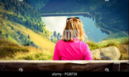 Eine blonde Frau, die alleine auf einer Bank sitzend in die Berge. Sie ist mit Blick auf ein Tal mit einem See. Vintage geschmack Farben Stockfoto