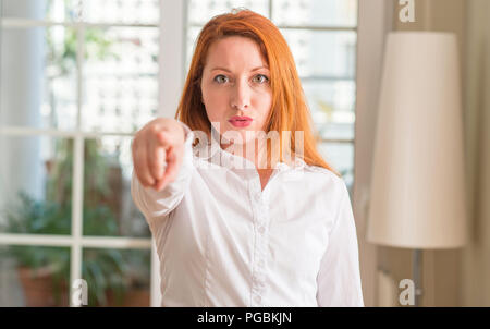 Rothaarige Frau mit weißem Hemd zu Hause mit dem Finger an die Kamera und an Sie gerichtet, Handzeichen, positiv und zuversichtlich Geste von vorne Stockfoto