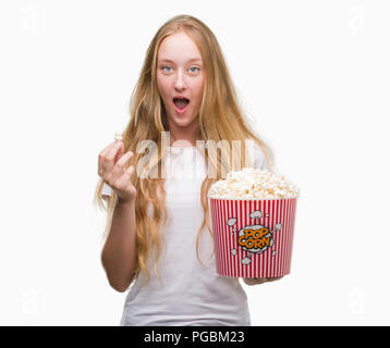 Blonde Teenager Frau essen Pop Corn Angst im Schlag mit einer Überraschung konfrontiert, ängstlich und aufgeregt mit Angst Ausdruck Stockfoto