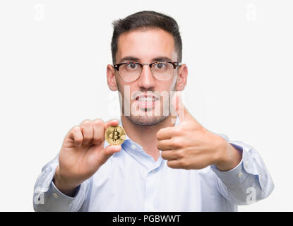 Hübscher junger Mann hält Bitcoin zufrieden mit Lächeln tun ok Zeichen, Daumen hoch, mit den Fingern, ausgezeichnete Zeichen Stockfoto