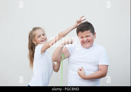 Lustige kaukasischen dünne kleine Mädchen messen lächelnd Fat Boy Muskel durch Band durchgeklickt Stockfoto