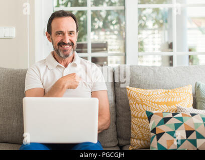 Mittleres alter Mann mit Laptop am Sofa mit Überraschung Gesicht zeigenden Finger zu sich selbst Stockfoto