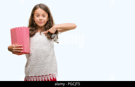 Brunette hispanische Mädchen essen Popcorn mit Überraschung Gesicht zeigenden Finger zu sich selbst Stockfoto