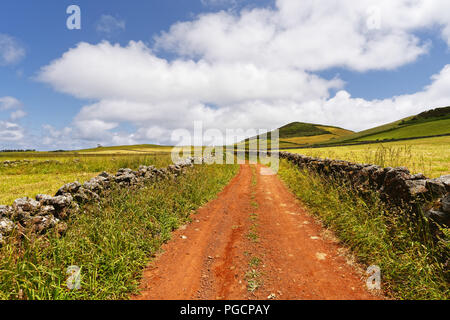 Eine rote Strecke führt durch eine hügelige Landschaft, am Rande des natürlichen Steinmauern, große Tiefe, Himmel mit einigen Wolken - Ort: Azoren, Insel Sao J Stockfoto