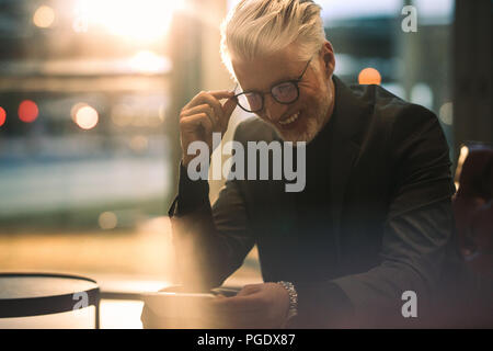 Gerne reife Geschäftsmann in Office Lobby sitzen und Ansehen von Videos auf Smart Phone. Mann in formelle Kleidung genießen Sie online Video auf seinem mobilen Stockfoto