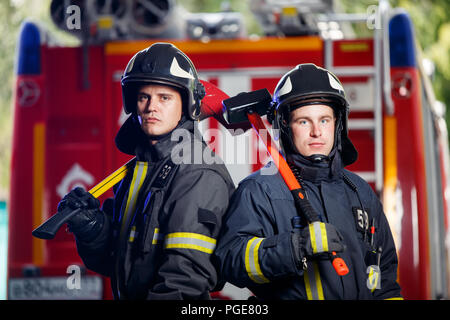 Foto von zwei Feuerwehrleuten Männer in Kamera mit Achsen in den Händen in der Nähe von Feuer Motor Stockfoto