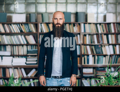 Portrait von bärtigen hipster Mann in der Bibliothek. Glatzköpfige Mann mit langem Bart steht auf dem Hintergrund der Bücher. Bildung Konzept Stockfoto
