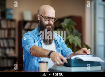 Bärtige stilvolle Schriftsteller schreiben auf der Schreibmaschine. Moderne Schriftsteller in Gläsern auf neues Buch arbeiten in der Bibliothek Stockfoto