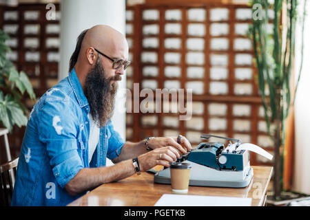 Bärtige stilvolle Schriftsteller schreiben auf der Schreibmaschine. Moderne Schriftsteller in Gläsern auf neues Buch arbeiten in der Bibliothek Stockfoto