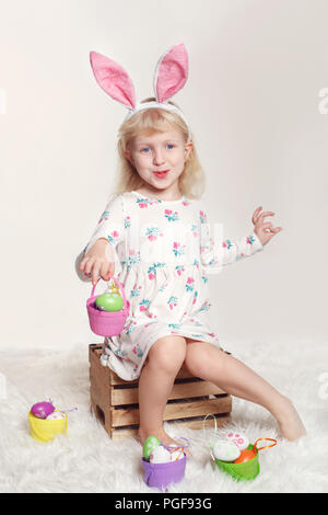 Cute adorable Kaukasier Kind Mädchen mit Osterhase hase Ohren sitzen auf Holzkiste im Studio. Kid holding Urlaub Korb mit bunten Eiern Cel Stockfoto