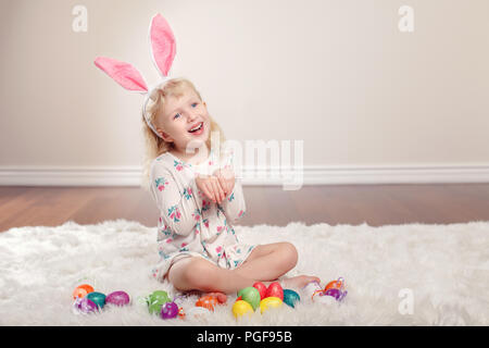 Cute adorable lustig Kaukasier Kind Mädchen mit Osterhase hase Ohren sitzen auf weichen, flauschigen Teppich Teppich im Studio. Kind Urlaub mit bunten Eiern Stockfoto