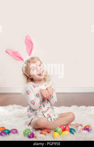 Cute adorable lustig Kaukasier Kind Mädchen mit Osterhase hase Ohren sitzen auf weichen, flauschigen Teppich Teppich im Studio. Kind Urlaub mit bunten Eiern Stockfoto