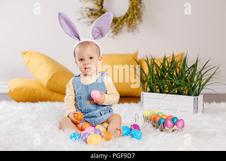 Cute adorable kaukasische Baby Mädchen in Jeans Kleid tragen Purple bunny Ohren sitzen im Studio. Kid Kind spielen mit Ostern bunte Eier feiern Stockfoto