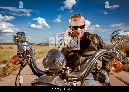 Biker sprechen auf einem Smartphone. Biker Mann mit einer Lederjacke und Sonnenbrille sitzt auf seinem Motorrad. Stockfoto