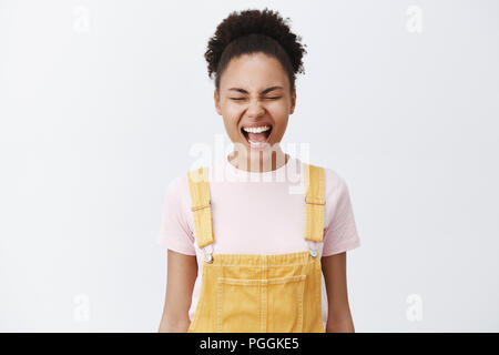 Gefühle lassen sich gut fühlt. Unvorsichtige schöne afrikanische Amerikaner mit glatten und sauberen Haut gelb Overalls, schreien, mit geschlossenen Augen, lösen Stress, die versuchen, beruhigen Sie sich über graue Wand Stockfoto