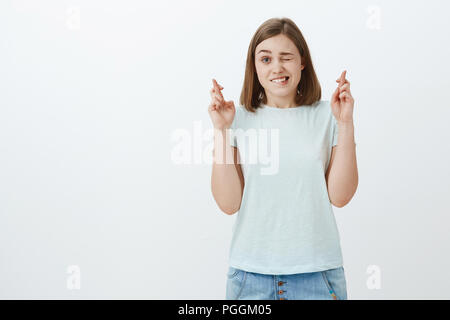 Studio shot von niedlichen aufgeregt und hoffnungsvolle weibliche Fan beobachten wichtige Spiel peeking mit einem Auge und drückt die Daumen für gutes Glück nervös Beobachten wichtige Spiel der Lieblingsmannschaft Stockfoto