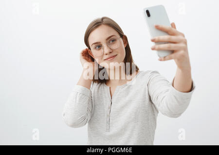 Smart und zärtliche Freundin in Gläser flippen Haarsträhne hinter dem Ohr und lächelte süß während der Einnahme von selfie auf neues Smartphone Posing über grauer Hintergrund erfreut die Post im sozialen Netz Stockfoto