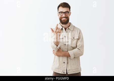 Zufrieden zufrieden charmante bärtigen Kerl mit trendigen Frisur, Brille in schwarz und beige Outdoor Jacke zeigt mit dem Zeigefinger auf Kamera, als ob näher kommen, einladenden Lächeln tricky und zuversichtlich Stockfoto