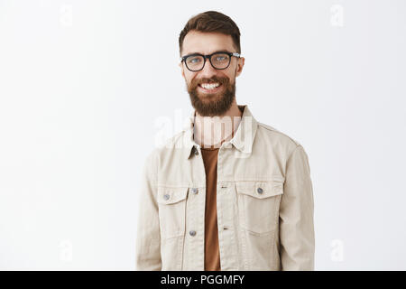 Taille - geschossen von freundlich - auf der Suche zufrieden stattlichen kaukasischen bärtigen Mann mit Schnurrbart lächelte zufrieden und glücklich Ständigen freudig über grauer Hintergrund in beige Jacke Stockfoto