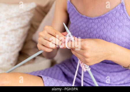 Nahaufnahme der Frau in einem lila Home Kleid strickt Ein hellrosa Schal oder Plaid aus natürlichen Threads auf dem Bett. Close-up horizontale Foto. Freiberufliche creati Stockfoto