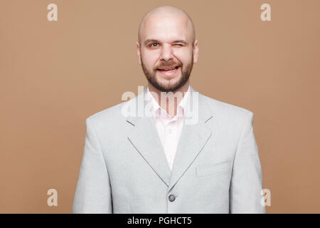 Portrait von Happy lustig aussehenden im mittleren Alter kahl bärtigen Geschäftsmann in Classic Light grauen Anzug stehen und zwinkerte Kamera schaut. indoor Studio Stockfoto
