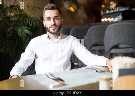 Portrait von gut aussehenden bärtigen Geschäftsmann tragen weisse Shirt sitzen am Tisch im Restaurant und an der Kamera zuversichtlich, kopieren Raum Stockfoto