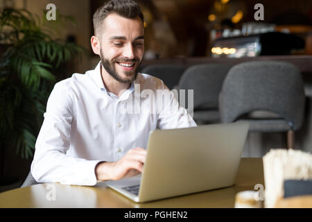 Portrait von gut aussehenden bärtigen Geschäftsmann tragen weisse Shirt glücklich lächelnd, während mit Laptop am Tisch im Cafe sitzen, Platz kopieren Stockfoto