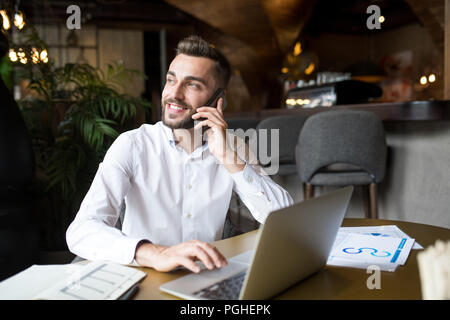 Portrait der junge Bartgeier Geschäftsmann sprechen durch Telefon und lächelt glücklich beim Arbeiten mit Laptop im Cafe Stockfoto