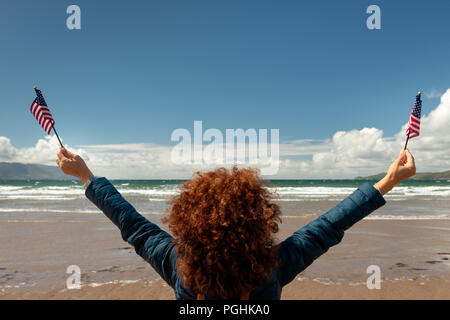 Ansicht der Rückseite des junge Frau mit roten lockigen Haar mit Blick auf das Meer, während die amerikanische Flaggen an einem Sandstrand auf windigen sonniger Tag mit blauem Himmel. Stockfoto