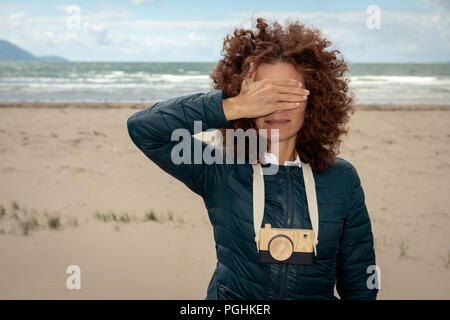 Junge, wunderschöne Frau mit roten Locken, die mit einer hölzernen Spielzeug-Fotokamera am leeren Strand posiert und dabei ihre Augen mit der Hand versteckt Stockfoto