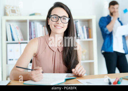 Porträt der hübsche junge Frau Brille fröhlich lächelnd, während sie Arbeiten am Schreibtisch im Büro oder in der Hochschule Stockfoto