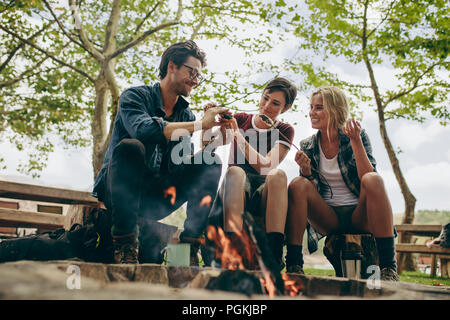 Lächelnd Freunde im Urlaub essen am Lagerfeuer toasten. Freunde machen ein Sandwich mit gerösteten Marshmallows sitzen draußen in der Landschaft. Stockfoto