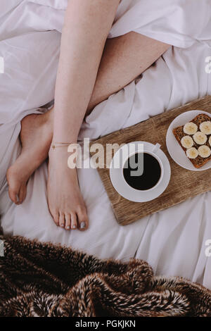 Nahaufnahme Beine einer Frau schlafen im Bett mit Frühstück durch ihre Seite. Frühstück mit Kaffee schwarz platziert neben eine Frau schlafend auf dem Bett. Stockfoto