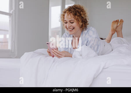 Glückliche Frau liegend auf Bett prüfen ihr Handy. Frau mit Ihrem Mobiltelefon im Bett nach dem Aufwachen am Morgen. Stockfoto